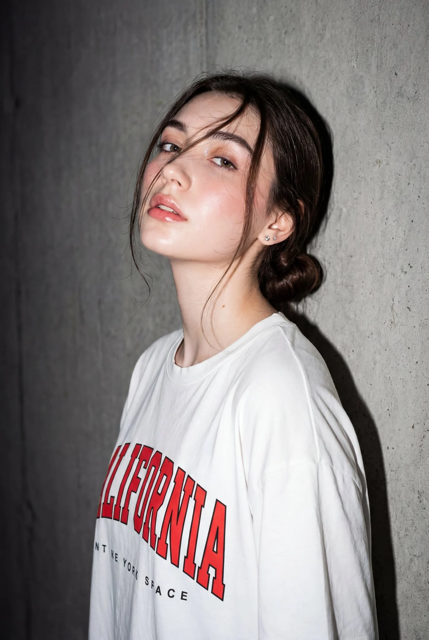 A striking editorial-style portrait of a young woman against a gray concrete wall, illuminated by direct flash, wearing a white 'CALIFORNIA' t-shirt.