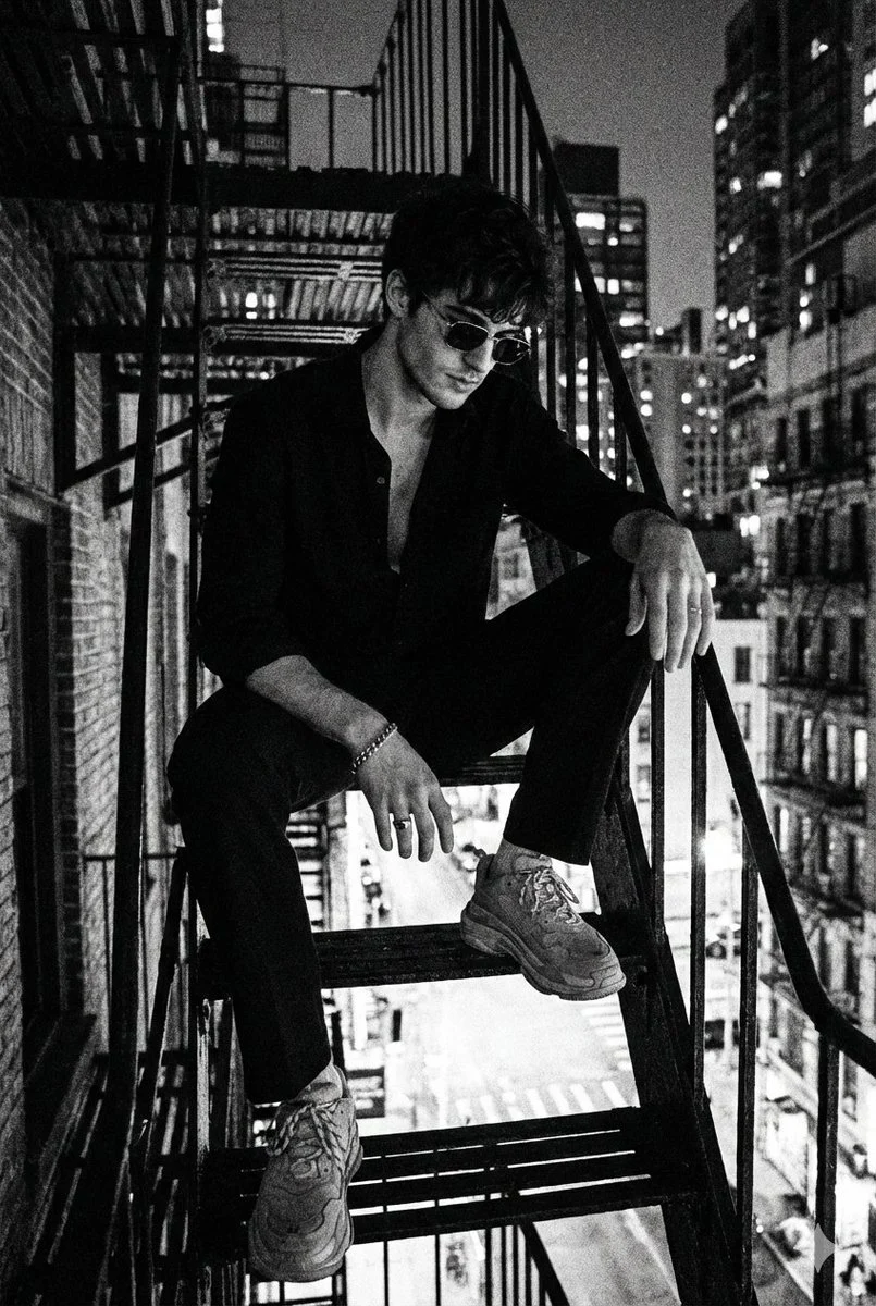 A high-contrast black and white noir man staircase portrait, featuring a man in dark attire on a metal staircase with a blurred cityscape background, exhibiting 90s grunge aesthetics and film grain.