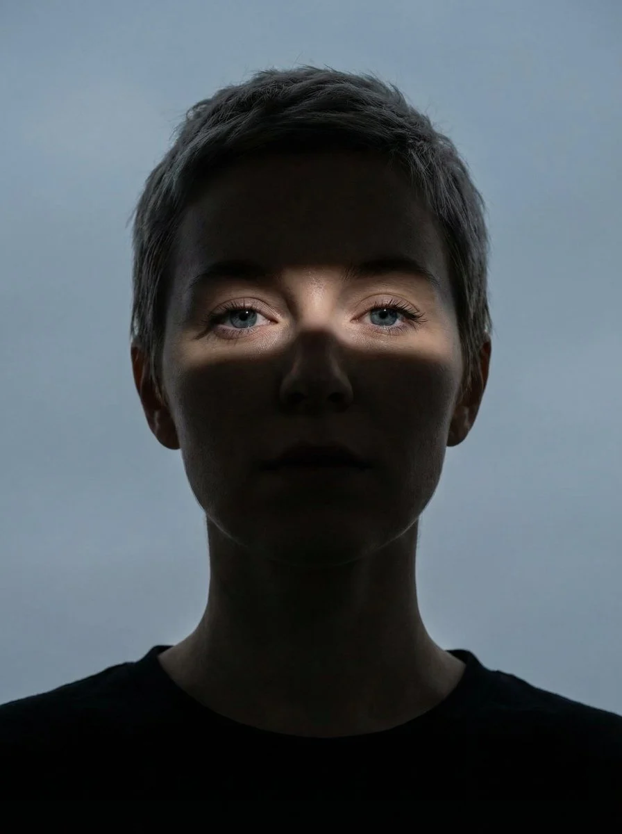 A striking close-up portrait of a woman with her face mostly in shadow, illuminated by a narrow horizontal light band across her eyes and nose, set against a plain grayish-blue sky.