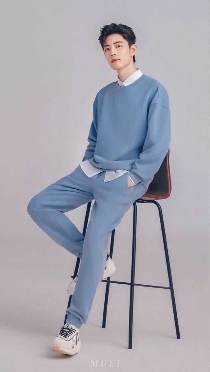 A stylish young East Asian man in a light blue oversized sweatshirt and joggers, leaning against a black barstool in a minimalist studio, embodying a Korean fashion studio man portrait aesthetic. - Variant 2